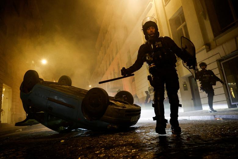 Các sĩ quan cảnh sát chống bạo động Pháp đi bên cạnh một chiếc xe bị lật ngửa trong cuộc biểu tình. (Ảnh: REUTERS/Juan Medina)