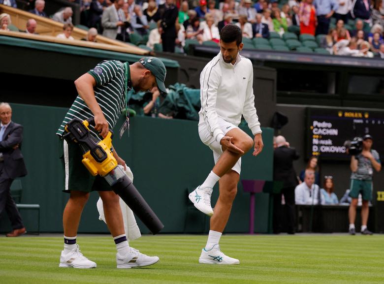 Novak Djokovic của Serbia cùng với một nhân viên mặt đất cố gắng làm khô sân sau cơn mưa trong trận đấu vòng đầu tiên với Pedro Cachin của Argentina tại Wimbledon (London, Anh). (Ảnh: REUTERS/Andrew Canridge)