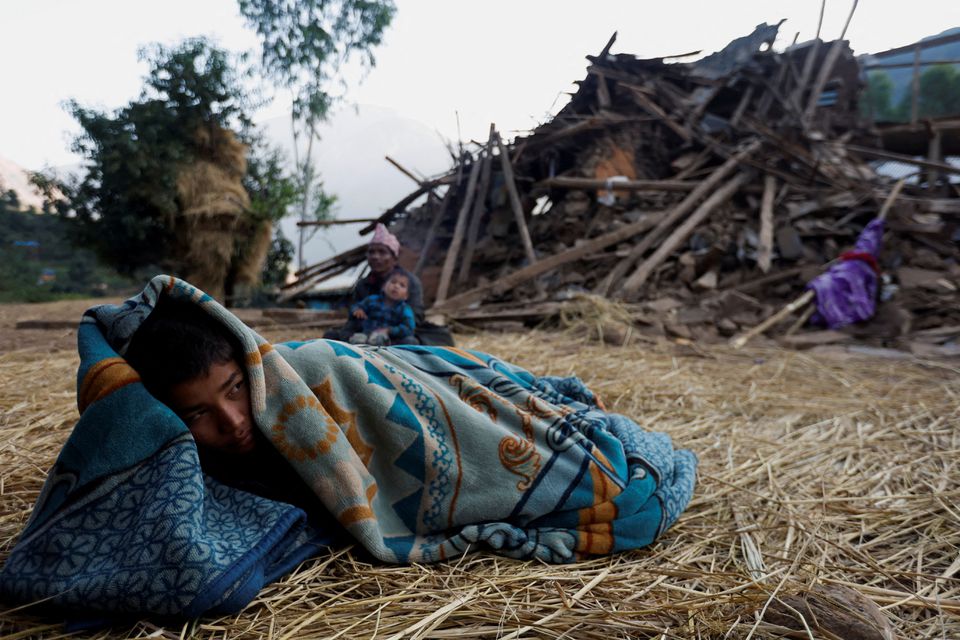 Một cậu bé đắp chăn nằm cạnh một ngôi nhà bị sập trong trận động đất ở Jajarkot, Nepal. (Ảnh: REUTERS)