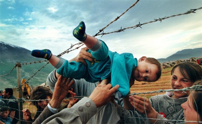 Một cậu bé hai tuổi, con trai của những người tị nạn chạy trốn khỏi xung đột, được chuyển qua một hàng rào dây thép gai để an toàn trong cuộc chiến ở Kosovo