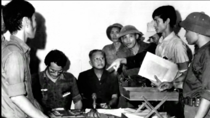 Bức ảnh giây phút lịch sử Dương Văn Minh tuyên bố đầu hàng tại đài phát thanh TP.HCM trưa 30/4/1975: Dương Văn Minh ngồi, mặc áo màu đen, Nguyễn Hữu Thái là người đứng, áo ngắn tay trắng, tay cầm xấp tài liệu. Ảnh: Tư liệu.