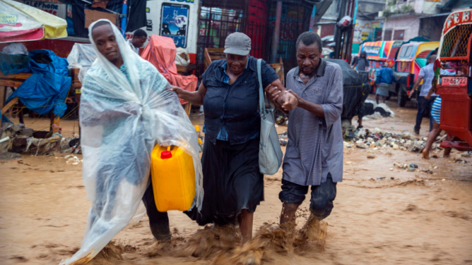 Người bán hàng rong giúp nhau đi qua đoạn đường nước chảy xiết do hoàn lưu bão ở Port-au-Prince, thủ đô Haiti. Ảnh: AP.