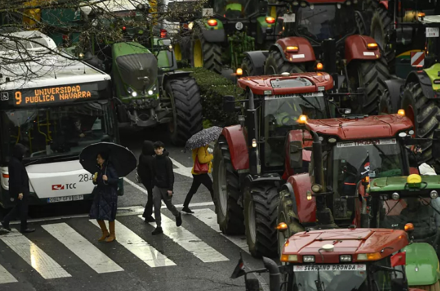 Pedestrian cross the road as farmers with their tractors march along the center of the city while taking part in a protest, in Pamplona, February 2024.