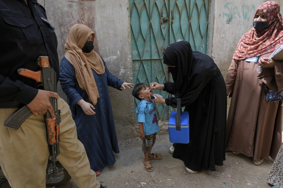 Cảnh sát đứng gác trong khi nhân viên y tế cho một đứa trẻ ở Karachi, Pakistan uống vaccine bại liệt. Ảnh: Fareed Khan.