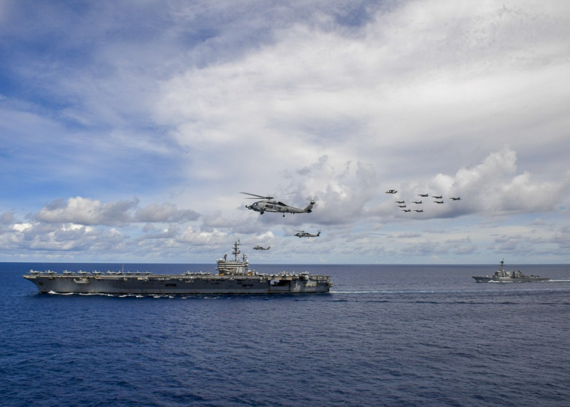 Ba trực thăng MH-60 bay gần tàu sân bay Reagan, phía trên cao là đội hình tiêm kích F/A-18 E/F Super Hornet, hộ tống phía sau là tàu khu trục lớp Arleigh Burke và tuần dương hạm lớp Ticonderoga