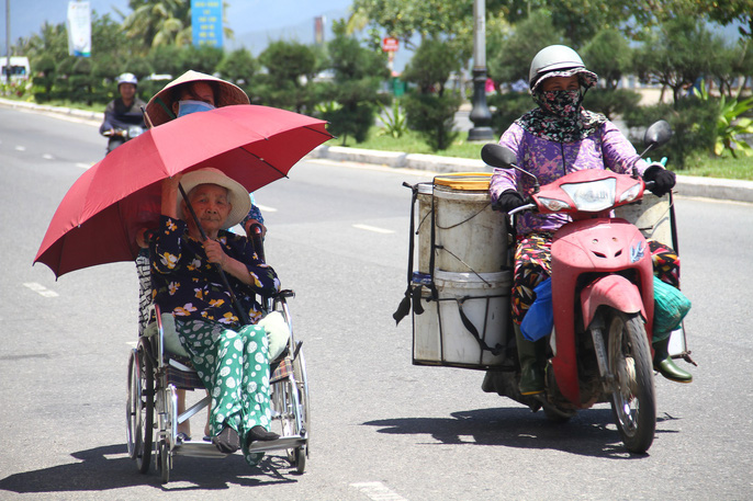 Một cụ già phải mang theo ô che nắng khi được người con gái đẩy xe lăn tại đường Nguyễn Tất Thành