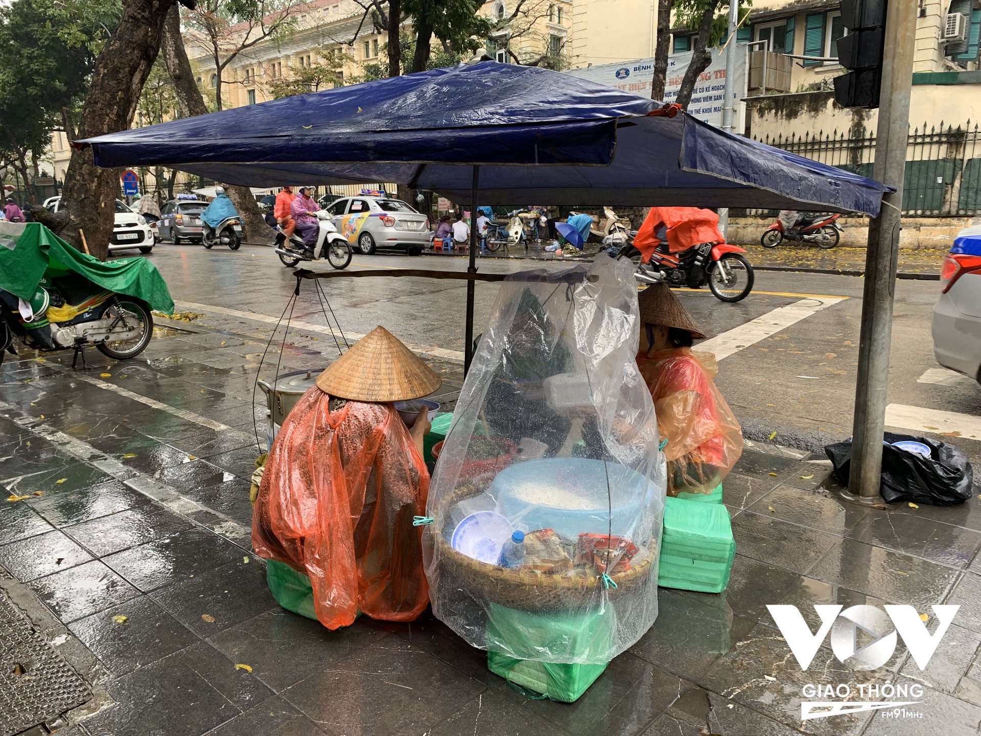 Một gánh hàng rong phục vụ ăn trưa trên vỉa hè phố Trần Bình Trọng, cả 'chủ quán' và khách đều chịu khó mặc áo mưa ngồi ăn giữa lúc trời mưa phùn rét buốt