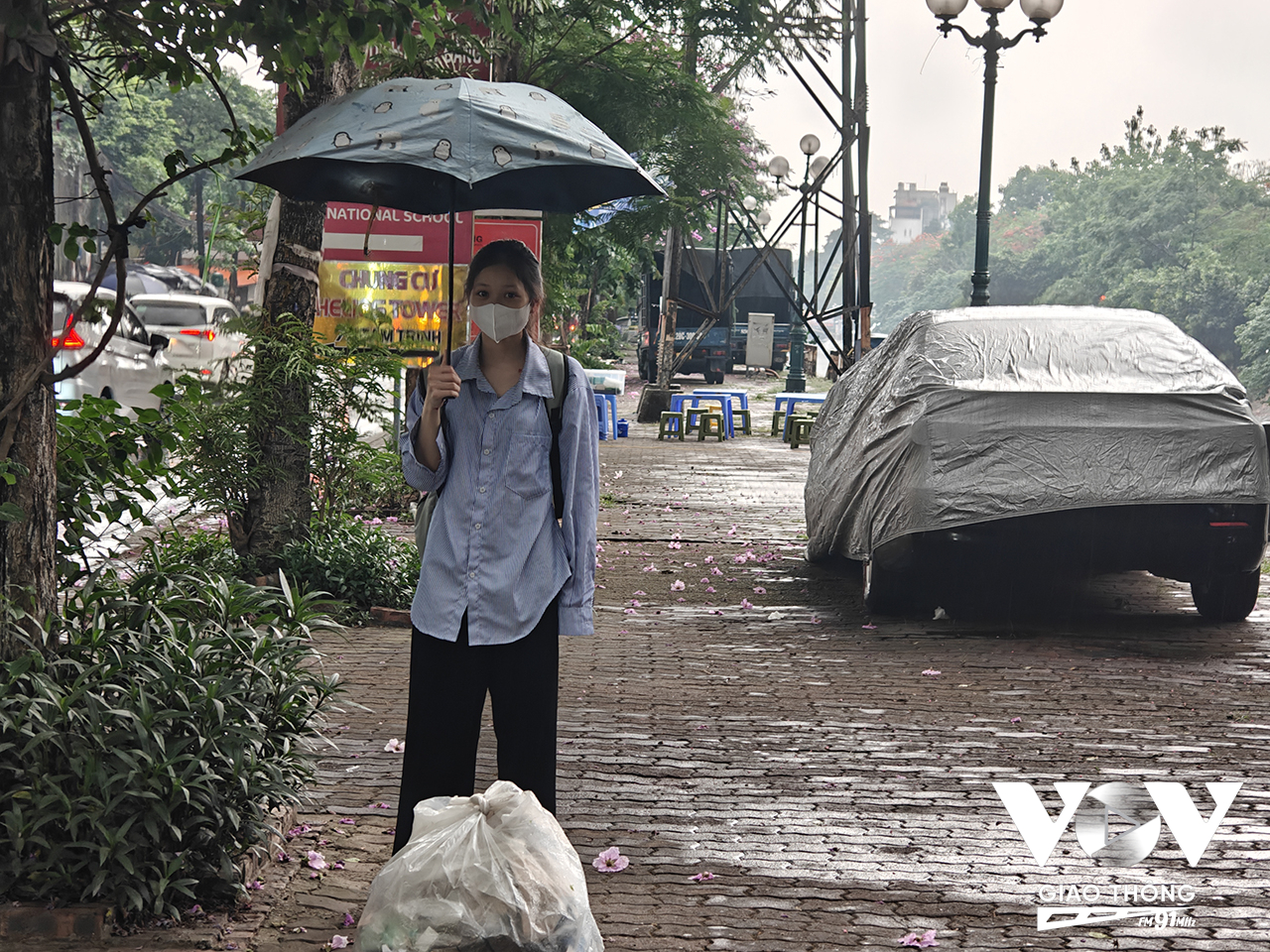 Phan Thị Thảo Mai, sinh viên năm ba, đang đứng đợi xe buýt bên cạnh rác thải, xe hơi và đằng xa là một quán nước phủ kín bề rộng vỉa hè