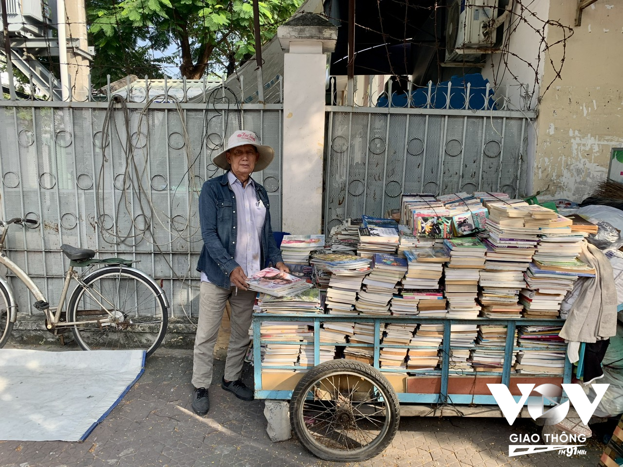 Ông cụ vẫn ngủ ven đường, “tổ ấm” nhỏ là chiếc võng cột tạm bên hàng cây, “gia tài” vỏn vẹn chỉ có chiếc xe đạp cũ kỹ cùng chiếc xe đẩy chứa hàng trăm quyển sách, báo cũ được được ông xếp ngay ngắn, tỷ mỉ.