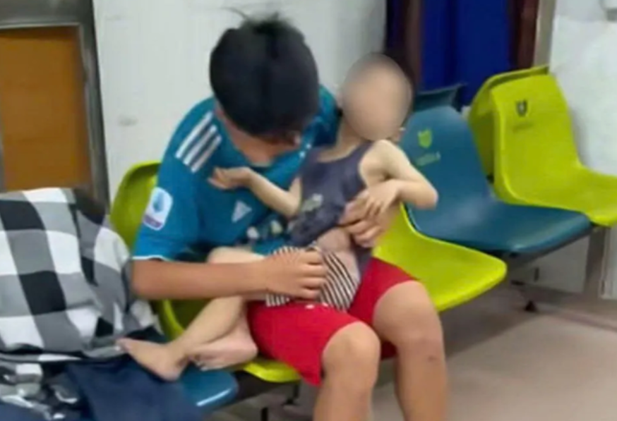 Bệnh nhi được bé trai lớn hơn bế ngồi ngoài hành lang Bệnh viện Đa khoa tỉnh Nam Định - Ảnh: Báo Tuổi trẻ